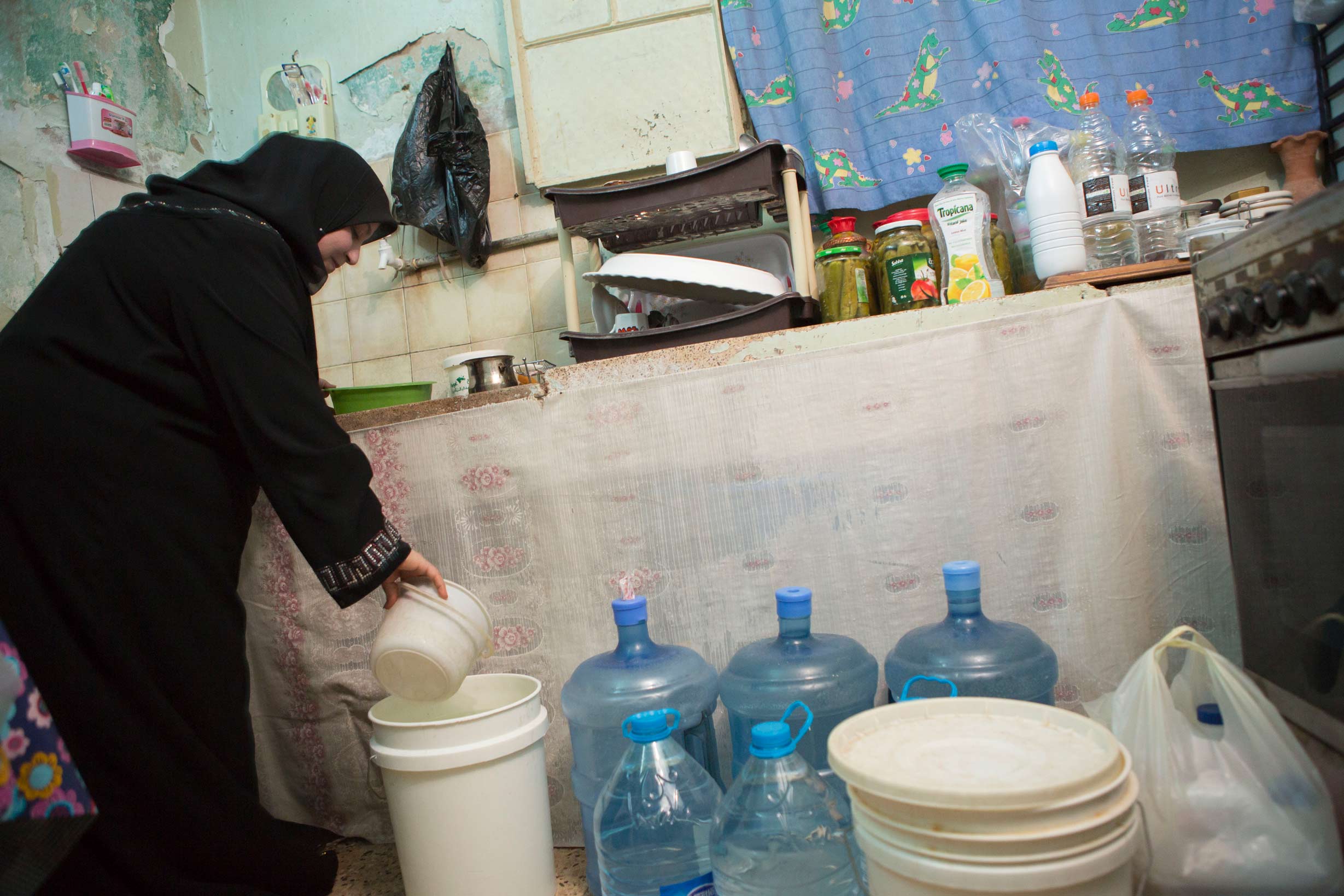 Mrs. Um Yazan Omran, a resident of Zarqa, Jordan, experiences water shortages. Here she is seen in her kitchen with several jugs of purchased water which she uses for the household.