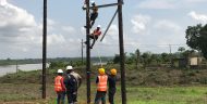 Two lineworkers on the ground hold a rope and two others are watching.