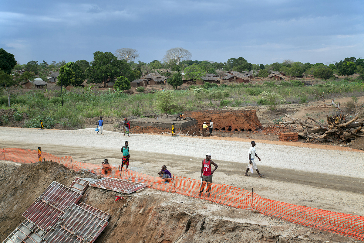 A road in Mozambique undergoing rehabilitation. There is a village in the distance and a road cutting across the image in the foreground, with stacks of bricks behind the road, and orange construction netting acting as a barrier in front. There are about a dozen people scattered about, including construction workers near the bricks, and some other people walking along the road.