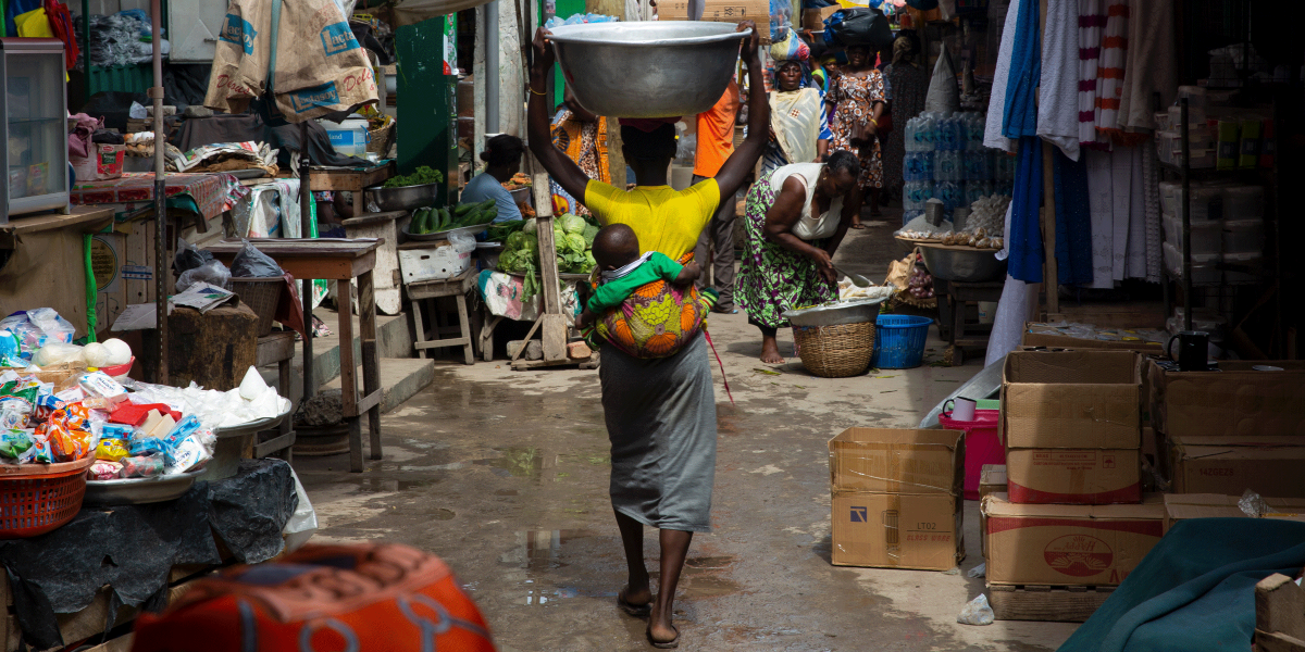 A woman with a baby on her back balances a basin on her head as she walks through a market in Ghana.